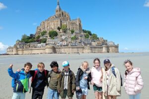 EIFA Huit enfants se tiennent pieds nus sur une plage de sable, avec le Mont-Saint-Michel, une abbaye insulaire historique en France, qui se dresse derrière eux sous un ciel bleu éclatant parsemé de nuages.