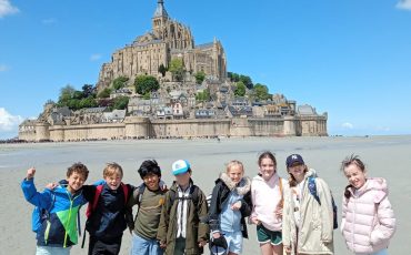 EIFA A group of eight children stand barefoot on the sandy beach in front of Mont Saint-Michel in France, smiling and posing for the photo on a sunny day with a blue sky and scattered clouds.