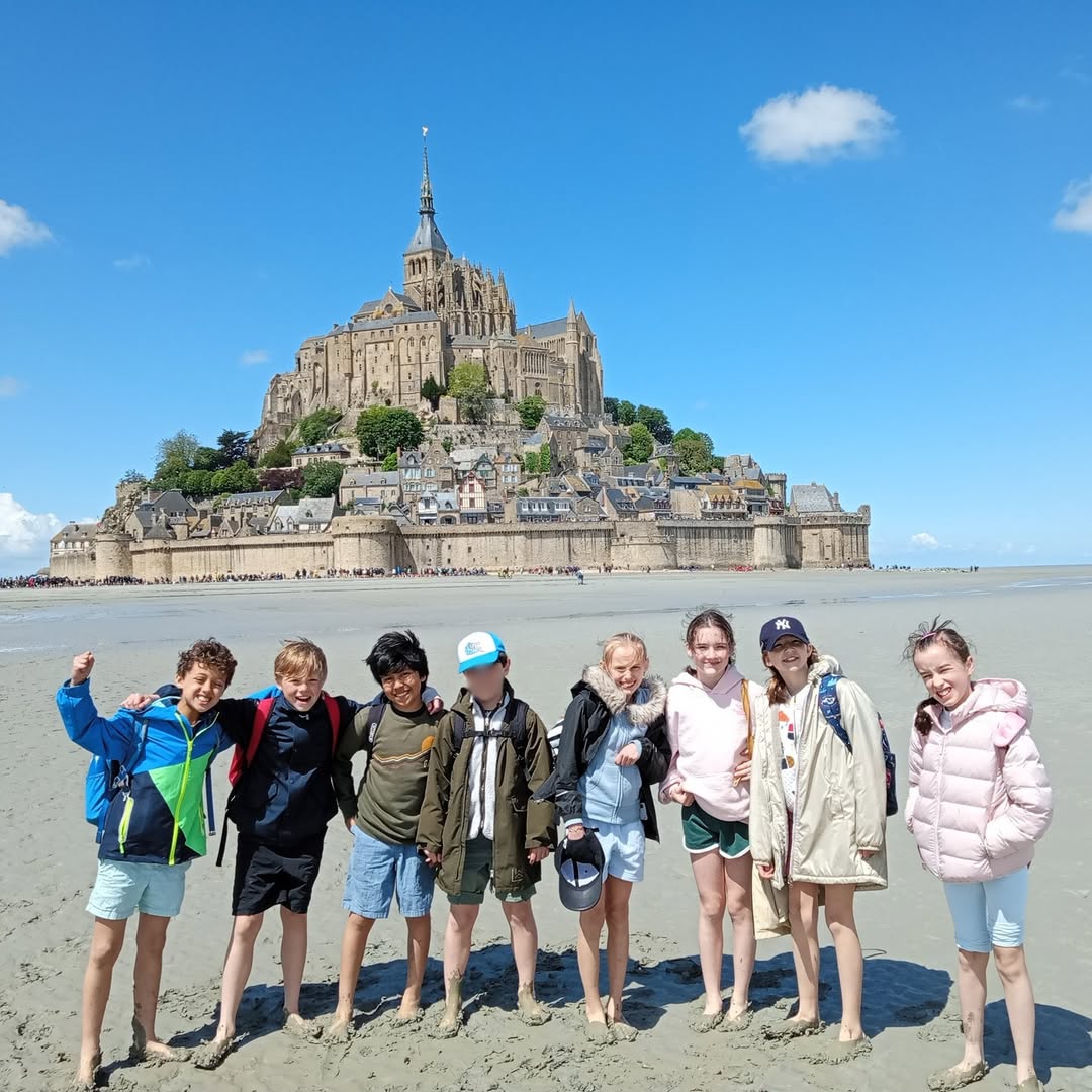 EIFA A group of eight children stand barefoot on the sandy beach in front of Mont Saint-Michel in France, smiling and posing for the photo on a sunny day with a blue sky and scattered clouds.