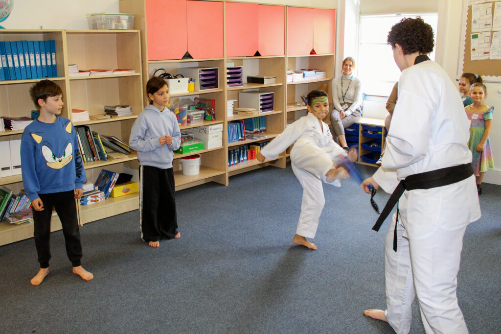 EIFA Children watch as a boy in a karate uniform kicks toward an instructor in a white belt and black belt. Shelves with books and supplies line the wall, and several adults and children observe the activity.