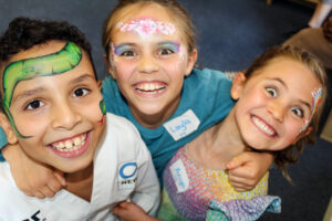 EIFA Trois enfants souriants, au visage peint de couleurs vives, posent ensemble pour célébrer la Journée mondiale de l'alimentation. L'un d'eux porte un uniforme d'arts martiaux, tandis que les autres portent des vêtements décontractés et des robes, leurs badges étant visibles alors qu'ils rayonnent d'excitation.