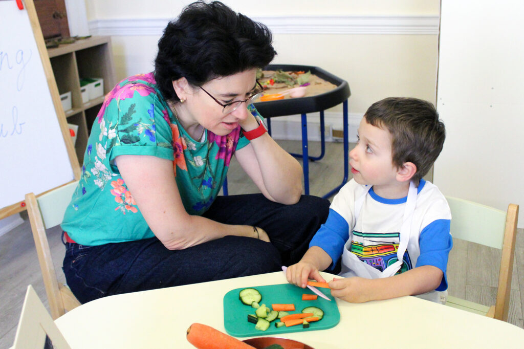 EIFA An adult woman and a young boy sit at a small table. The boy is holding a carrot stick, and there are assorted vegetables on a plate. They are looking at each other, engaged in conversation.
