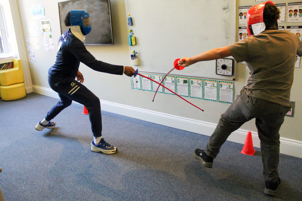 EIFA Two people in protective masks and casual clothes fence with plastic swords indoors, standing near a whiteboard and orange cones on a carpeted floor.
