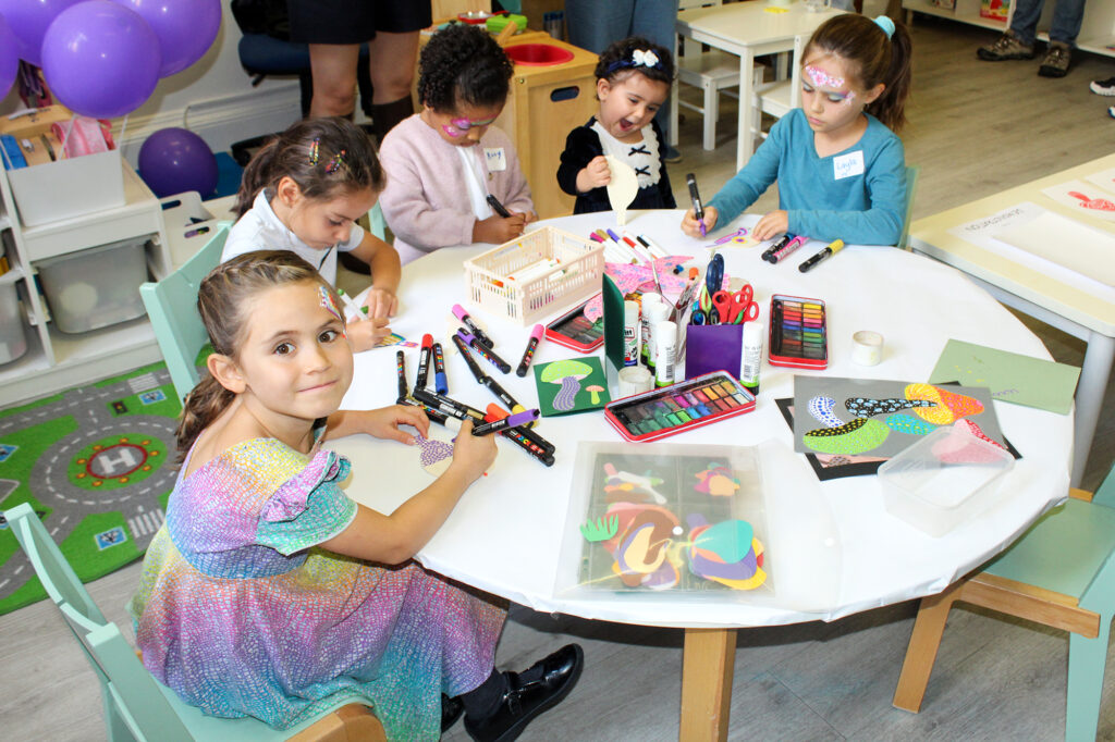 EIFA Five young children sit around a table doing arts and crafts with markers and paper. One girl in a sparkly dress looks at the camera, while the others are focused on their artwork. The setting is a colorful classroom.