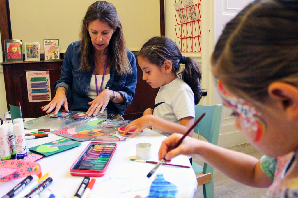 EIFA A woman and two young girls work on colorful art projects at a table covered with art supplies. One girl paints, while the other cuts paper. The woman appears to be guiding them. Artworks and supplies are visible in the background.