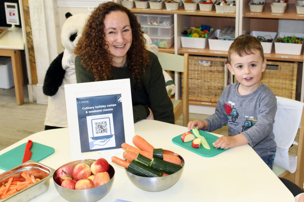 EIFA A woman and a young boy sit at a table with bowls of apples, carrots, and cucumbers. The boy is cutting vegetables, and a sign about culinary holiday camps with a QR code is on the table. A stuffed panda is in the background.