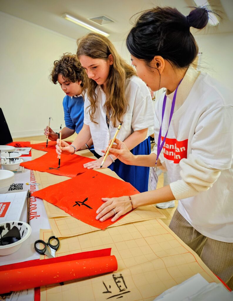 EIFA Three young people stand at a table practicing Chinese calligraphy, painting black characters on red paper with brushes. Supplies like ink, bowls, scissors, and paper are spread out on the table.