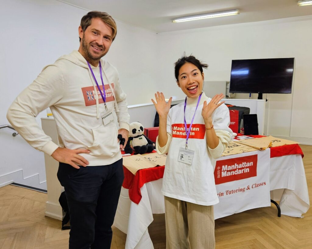 EIFA Two people wearing “Manhattan Mandarin” shirts and name badges smile at a booth with a red and white tablecloth, a panda plush, and learning materials laid out. The background is a bright room with wood flooring.