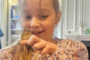 EIFA Une jeune fille aux longs cheveux blonds sourit en regardant un papillon perché sur son doigt. Elle porte une robe à motifs floraux et se trouve à l'intérieur, célébrant la Journée mondiale de l'alimentation, avec des bibliothèques et une cheminée blanche derrière elle.