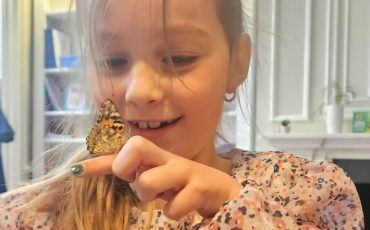 EIFA A young girl with blonde hair smiles as she gently holds a butterfly on her finger indoors. She wears a floral-patterned blouse and, during the International Open Day, looks at the butterfly with fascination. Bookshelves and a fireplace are in the background.