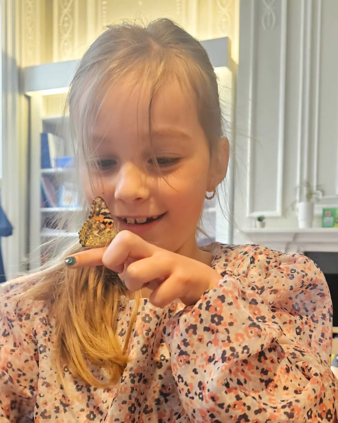 EIFA A young girl with blonde hair smiles as she gently holds a butterfly on her finger indoors. She wears a floral-patterned blouse and, during the International Open Day, looks at the butterfly with fascination. Bookshelves and a fireplace are in the background.