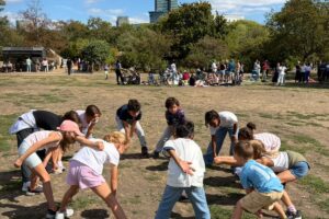 EIFA A group of children stand in a circle outdoors, bending forward and placing their hands on their knees, appearing to play a game on a sunny day with people and trees in the background.