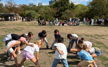 EIFA A group of children stand in a circle outdoors, bending forward and placing their hands on their knees, appearing to play a game on a sunny day with people and trees in the background.