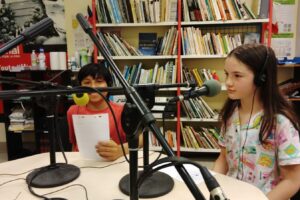 EIFA Two children wearing headsets sit at a table in a radio studio, holding papers and speaking into microphones. Shelves of books line the background, creating a lively and educational atmosphere.