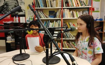 EIFA Two children wearing headsets sit at a table in a radio studio, holding papers and speaking into microphones. Shelves of books line the background, creating a lively and educational atmosphere.