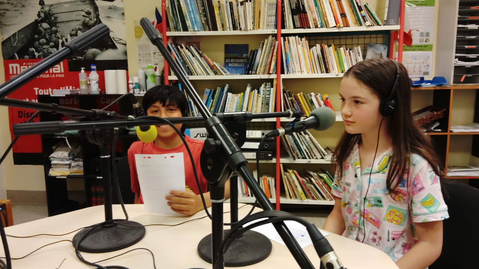EIFA Deux enfants, équipés d'écouteurs, sont assis à une table équipée de microphones, un document à la main, dans une pièce bordée d'étagères remplies de livres. On dirait qu'ils enregistrent ou interviennent dans le cadre d'une émission de radio ou d'un podcast.