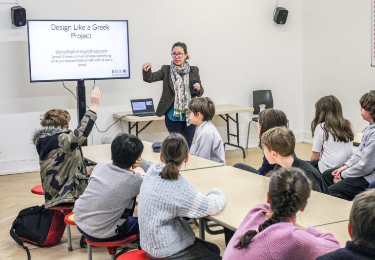 EIFA A teacher stands by a screen titled “Design Like a Greek Project,” speaking to a group of students seated at tables; one student raises their hand to participate.