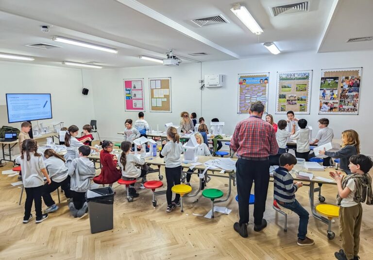 EIFA A classroom filled with children and a teacher working on a group project. Students are gathered around tables with papers and materials, some standing, some sitting on colorful stools, and others interacting in small groups.