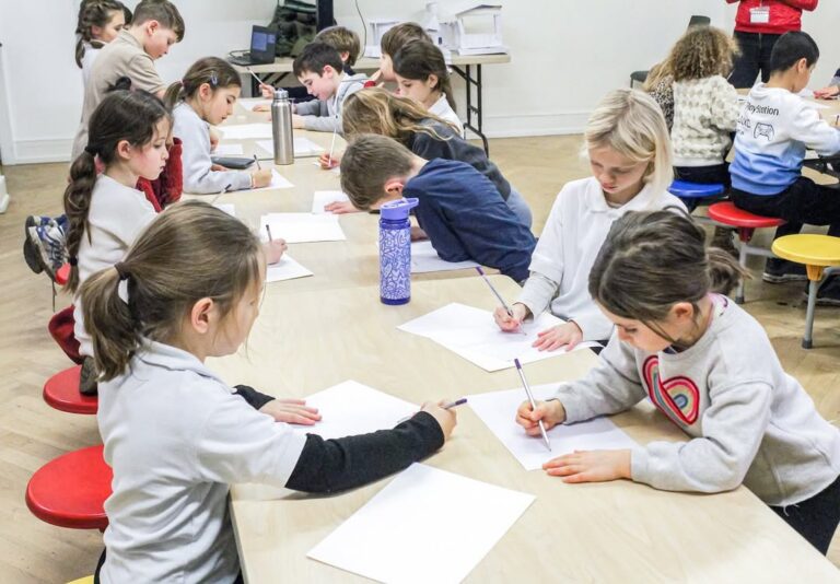 EIFA A group of young children sit around a table, focused on drawing or writing on sheets of paper with pencils. The classroom setting is bright, and an adult stands in the background.