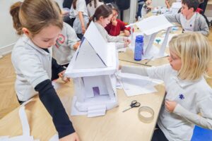 EIFA Dans une salle de classe, des enfants travaillent ensemble à la construction de maquettes de maisons en papier et en carton. Des ciseaux, du ruban adhésif et d’autres matériaux de bricolage sont éparpillés sur la table tandis qu’ils se concentrent sur leurs projets. EIFA Dans une salle de classe, des enfants travaillent ensemble à la construction de maquettes de maisons en papier et en carton. Des ciseaux, du ruban adhésif et d'autres matériaux de bricolage sont éparpillés sur la table tandis qu'ils se concentrent sur leurs projets.