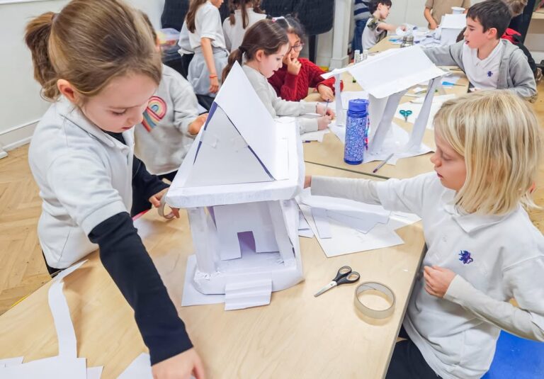 EIFA Children in a classroom work together around a table, building houses out of white paper and cardboard. Supplies like scissors, tape, and water bottles are scattered on the table. The atmosphere appears creative and collaborative.