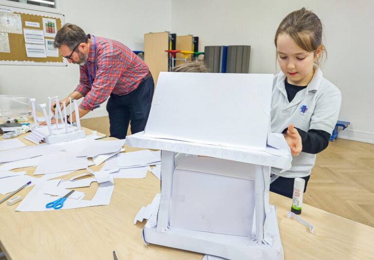 EIFA A young girl and an adult work together on a craft project, creating structures from white paper on a table covered with paper, scissors, and glue in a classroom setting.