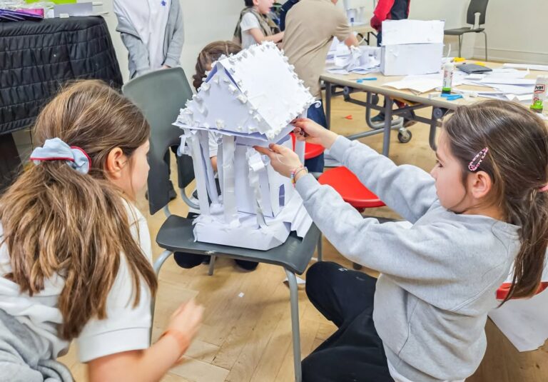 EIFA Two children work together to build a model structure out of white paper and card at a classroom table, while other students and paper projects are visible in the background.