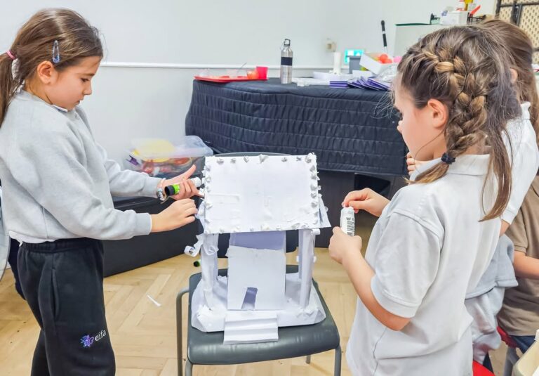 EIFA Two young girls are working together to decorate a model house made of white cardboard, using glue and small materials, in a classroom setting. The house is placed on a chair.