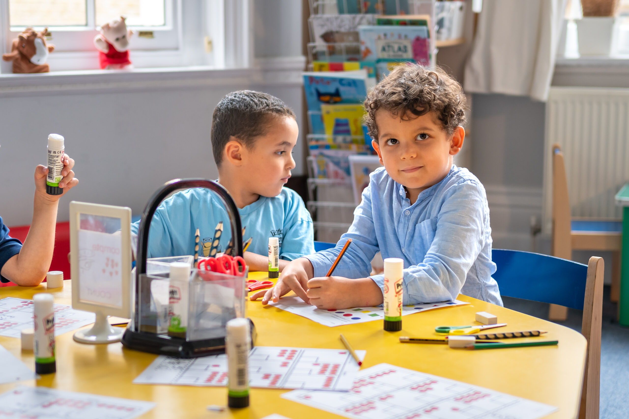 EIFA Deux jeunes garçons sont assis à une table jaune dans une salle de classe bilingue, travaillant sur des projets artisanaux et dessinant avec des bâtons de colle et des marqueurs. Une étagère avec des livres se trouve à larrière-plan près dune fenêtre, reflétant lenvironnement dapprentissage dynamique dune école EIFA.