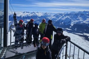 EIFA Un groupe de personnes en tenue de ski se tient sur un belvédère de montagne enneigée avec des skis, célébrant la Journée mondiale de l'alimentation sur fond de sommets enneigés et de ciel bleu clair.