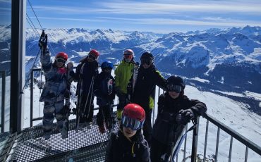 EIFA A group of people in ski gear stand on a snowy mountain platform during International Open Day, with a scenic backdrop of snow-covered peaks and a bright blue sky. Some hold skis, and everyone wears helmets and goggles.