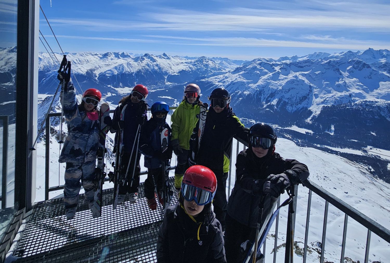 EIFA A group of people in ski gear stand on a snowy mountain platform during International Open Day, with a scenic backdrop of snow-covered peaks and a bright blue sky. Some hold skis, and everyone wears helmets and goggles.