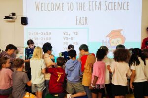 EIFA Un groupe d'enfants est rassemblé devant un panneau sur lequel on peut lire « Bienvenue au salon des sciences de l'EIFA ». Debout ou assis sur des tabourets colorés, ils tournent le dos à la caméra et regardent une table sur laquelle sont exposés des projets.