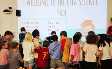 EIFA A group of children gathers in front of a large screen that reads “Welcome to the EIFA Science Fair” in a classroom, with scheduled times and categories displayed on the screen.