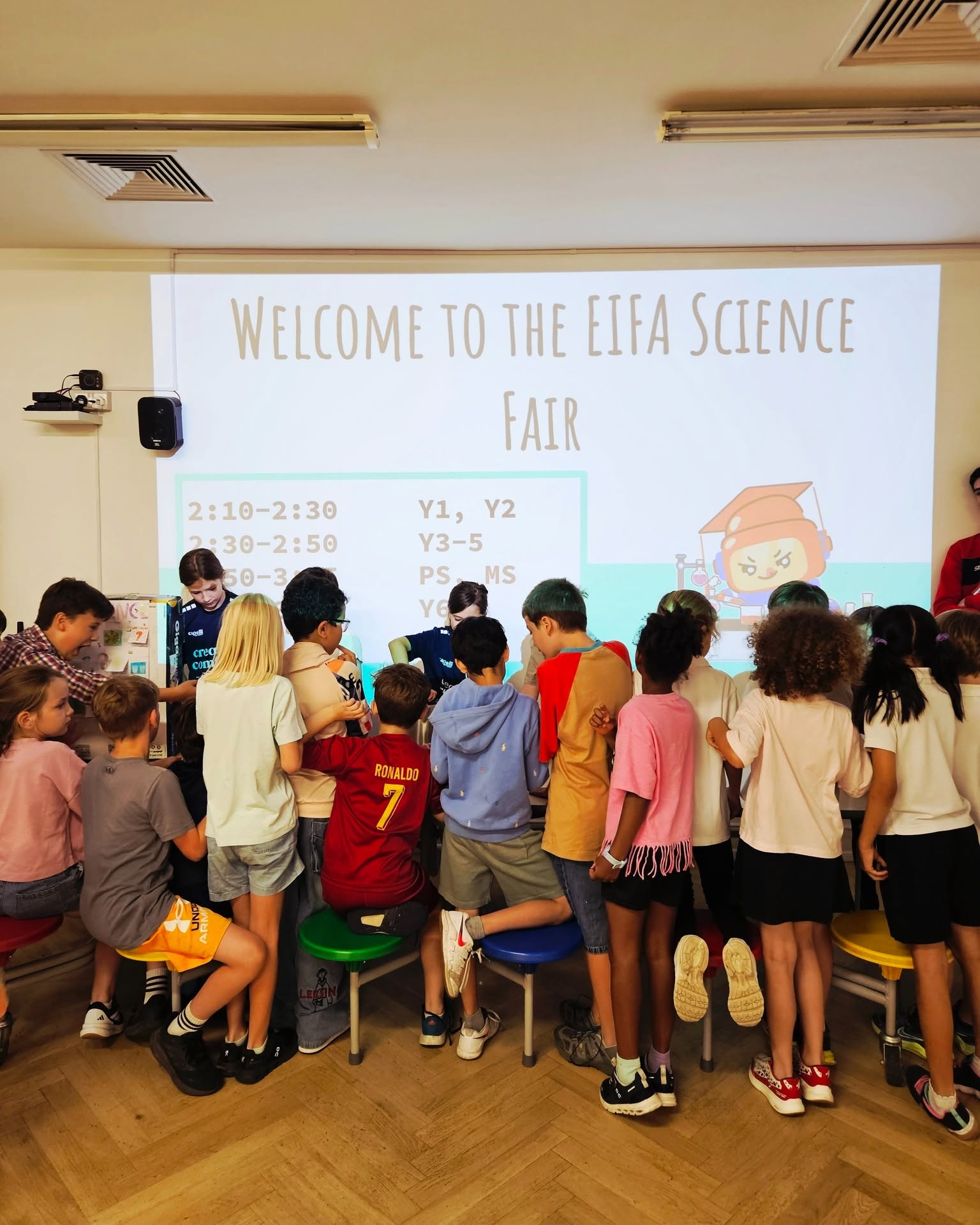 EIFA A group of children gathers in front of a large screen that reads “Welcome to the EIFA Science Fair” in a classroom, with scheduled times and categories displayed on the screen.
