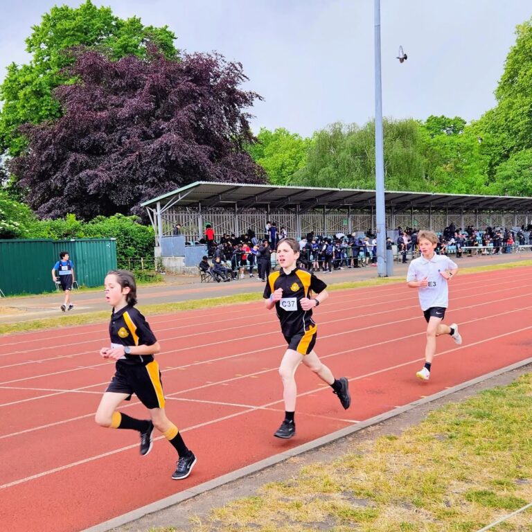 EIFA Trois jeunes garçons courent sur une piste d'athlétisme rouge lors d'une course organisée à l'occasion de la Journée mondiale de l'alimentation, avec des spectateurs dans une tribune couverte et des arbres verdoyants derrière eux. Deux garçons portent des uniformes noirs et jaunes, tandis qu'un autre est vêtu de blanc.