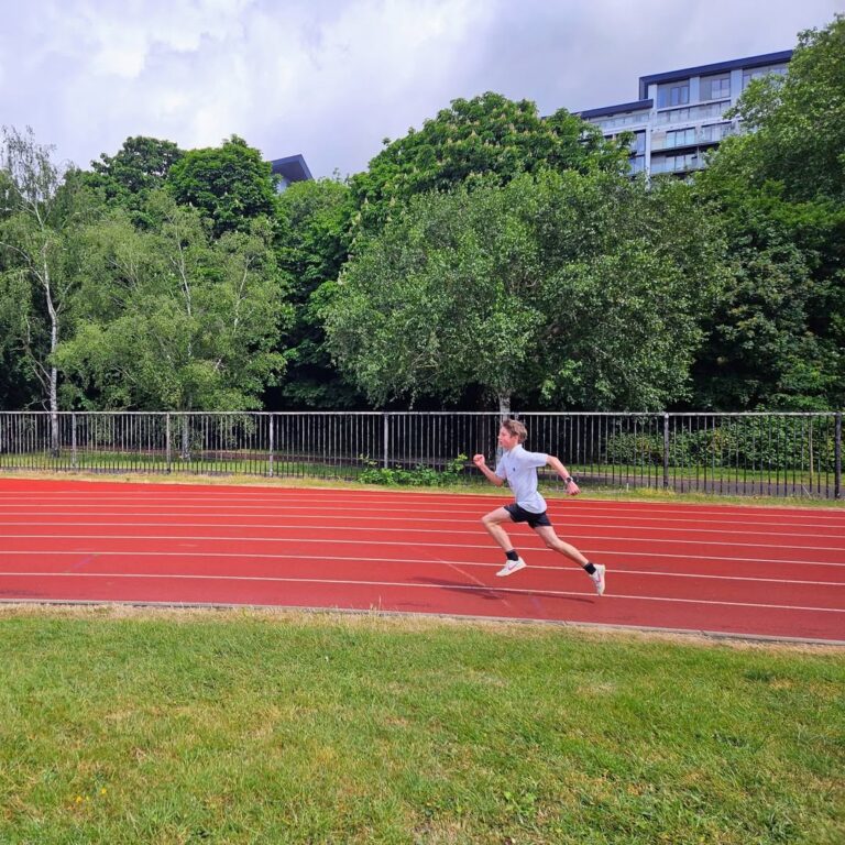 EIFA Une personne en tenue de sport sprinte sur une piste de course rouge en plein air, célébrant la Journée mondiale de l'alimentation, avec des arbres verts et un bâtiment visibles en arrière-plan sous un ciel partiellement nuageux.