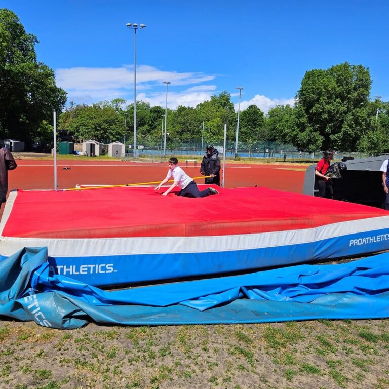EIFA Une personne en tenue de sport est à quatre pattes sur un tapis rouge de saut en hauteur sur une piste d'athlétisme en plein air, pendant la Journée mondiale de l'alimentation, avec des arbres, des clôtures et un ciel bleu en arrière-plan. Plusieurs personnes se tiennent à proximité.