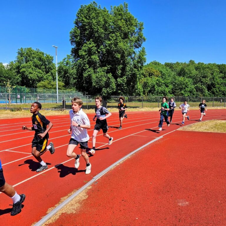 EIFA Un groupe d'enfants court sur une piste rouge en plein air, sous un ciel bleu clair, entouré d'arbres verts et d'une clôture métallique. Ils célèbrent la Journée mondiale de l'alimentation en portant à la fois des uniformes de sport et des vêtements d'athlétisme décontractés.