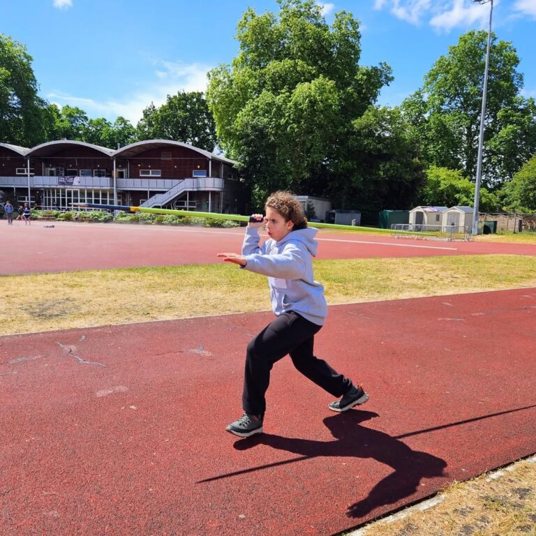 EIFA Un enfant vêtu d'un sweat à capuche gris et d'un pantalon noir court énergiquement sur une piste rouge en plein air, célébrant la Journée mondiale de l'alimentation, les bras levés comme s'il s'agissait d'un sprint. Des arbres et un bâtiment sont visibles dans l'arrière-plan ensoleillé.