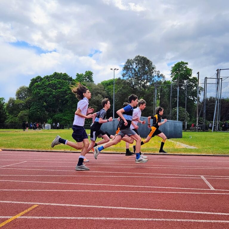 EIFA Cinq jeunes athlètes courent sur une piste extérieure, à mi-course, sous un ciel partiellement nuageux. Des arbres et des équipements sportifs sont visibles à l'arrière-plan, ce qui donne une impression de compétition et de mouvement à l'occasion de la Journée mondiale de l'alimentation.