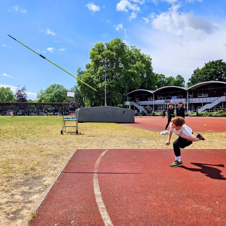 EIFA Une personne en plein mouvement lance un javelot sur une piste d'athlétisme en plein air pendant la Journée mondiale de l'alimentation, avec des arbres, des bâtiments et des spectateurs visibles à l'arrière-plan sous un ciel partiellement nuageux.