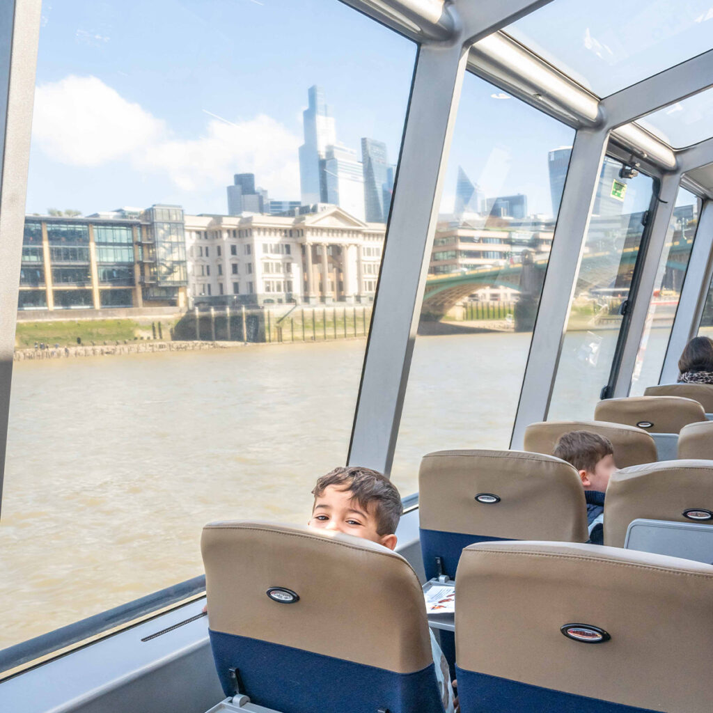 EIFA A young boy peeks over the back of a seat on a boat. Through the large windows, a cityscape and river are visible, including modern skyscrapers and historic buildings. Several other passengers are seen seated.