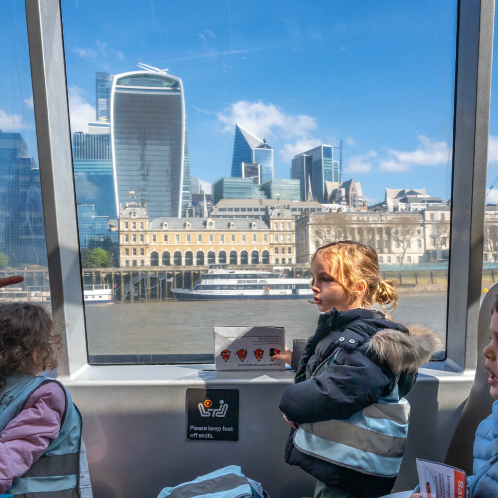 EIFA Two children wearing jackets sit near a window on a boat, with one holding a brochure. The window shows a view of modern skyscrapers and historic buildings along the Thames River in London.