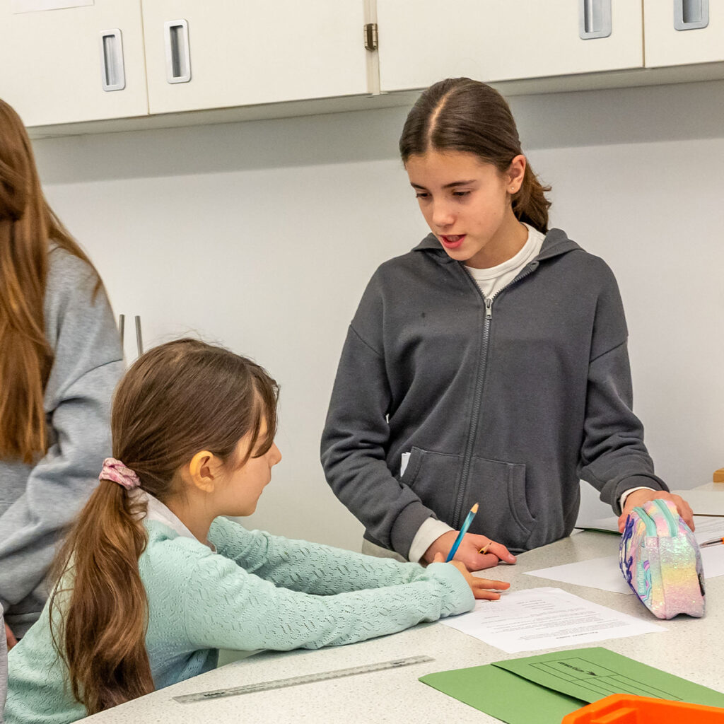 EIFA Two girls in a classroom are engaged in a discussion. One girl leans on a table holding a pencil, while the other stands, holding a pencil case. Papers and school supplies are on the table. Cabinets are visible in the background.