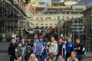 EIFA Un groupe d'enfants pose et sourit devant l'entrée du London Transport Museum. Les portes en verre derrière eux affichent le nom du musée et des lumières de fête sont visibles au-dessus. Les enfants portent des vestes et des vêtements décontractés.
