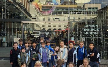 EIFA A group of children pose and smile in front of the entrance to the London Transport Museum. The glass doors behind them display the museums name, and festive lights are visible around the entrance.
