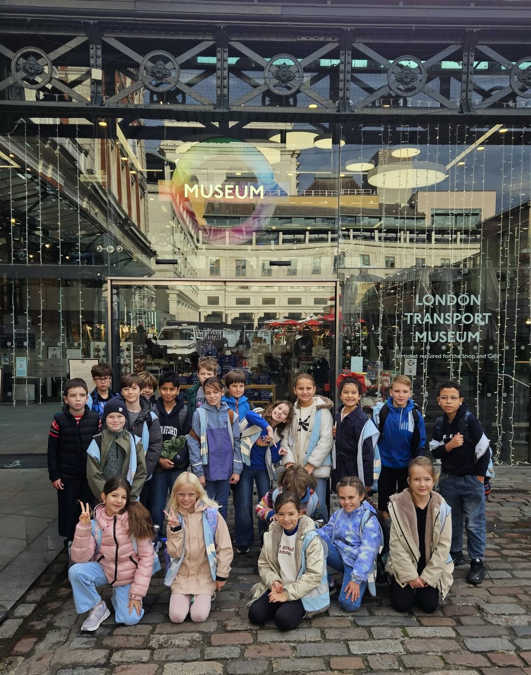 EIFA Un groupe d'enfants pose et sourit devant l'entrée du London Transport Museum. Les portes en verre derrière eux affichent le nom du musée et des lumières de fête sont visibles au-dessus. Les enfants portent des vestes et des vêtements décontractés.