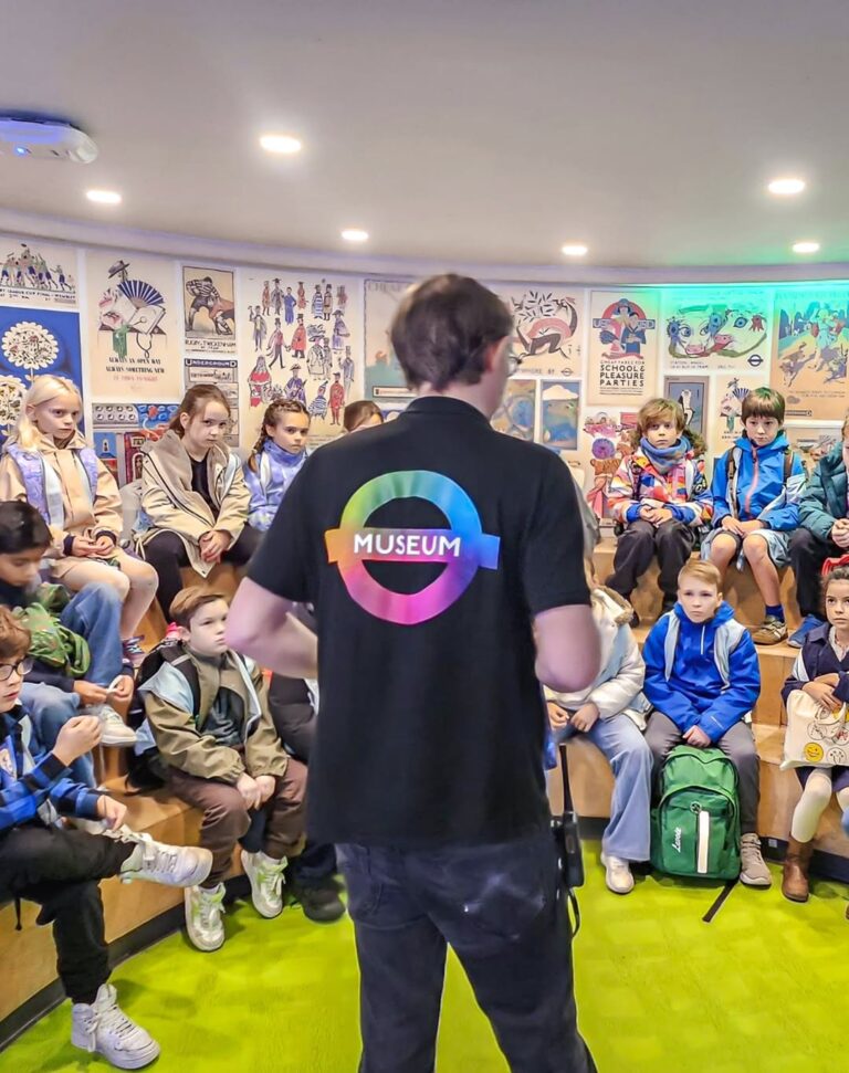 EIFA A man wearing a black shirt with a colorful “Museum” logo stands in front of a group of children seated on tiered benches, listening attentively in a brightly decorated, poster-filled room.