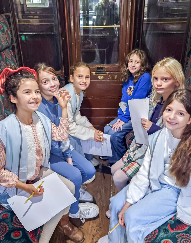 EIFA Six filles souriantes sont assises ensemble dans un compartiment de train ancien, tenant des papiers et des crayons, et regardant la caméra. Elles semblent être engagées dans une activité ou un projet scolaire.